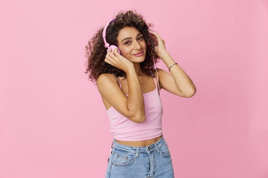 Happy woman wearing headphones with curly hair listening to music and singing along with her eyes closed in a pink T-shirt and jeans on a pink background, copy space. High quality photo