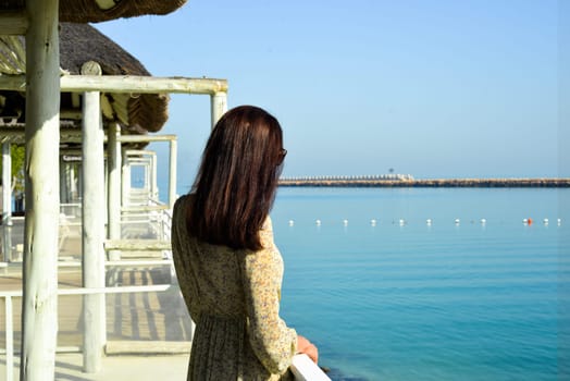 Young happy woman in a cute dress sits on a pier by the sea enjoying summer sunbeams.