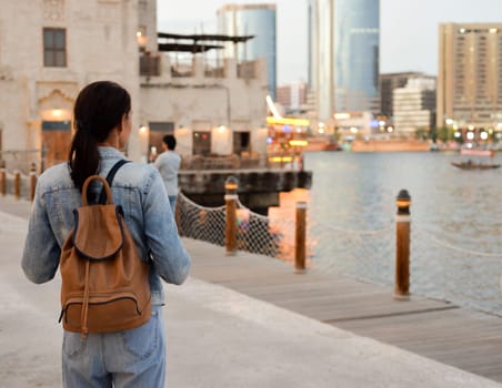 A young woman with a backpack on her back admires the river in the old Dubai Creek. Back view. Journey through the Persian Gulf.