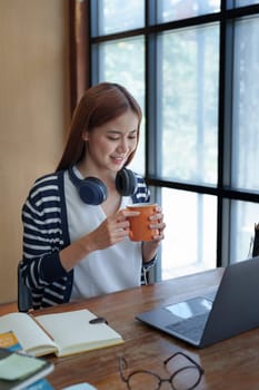 Portrait of a young, attractive Asian teenage girl using a computer and drinking coffee while studying online in the morning at the library.