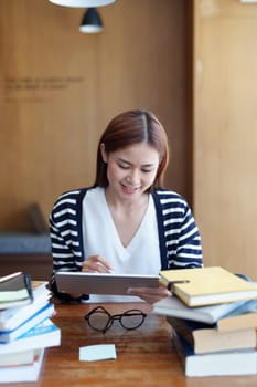 A portrait of a young Asian woman with a smiling face using a tablet computer during an online video conferencing class in a library.