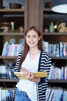 A portrait of a young Asian woman smiling while holding a notebook while researching in the library.