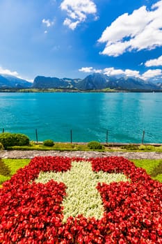 Flowerbed of the Swiss flag with boat cruise on the Thun lake and Alps mountains, Oberhofen, Switzerland. Swiss flag made of flowers and passenger cruise boat, Lake Thun, Switzerland.