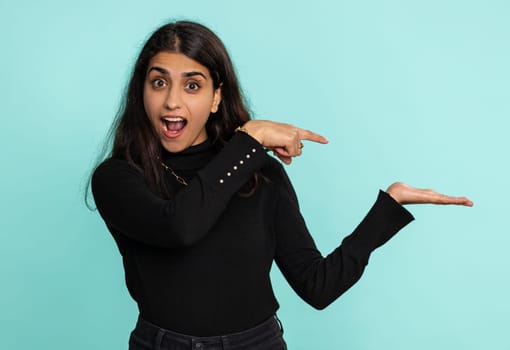 Young indian woman showing thumbs up and pointing empty place, advertising area for commercial text, copy space for goods promotion. Arabian hindu girl isolated alone on blue studio background indoors