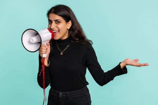 Indian woman talking with megaphone, proclaiming news, loudly announcing advertisement, warning using loudspeaker to shout speech, pointing empty place. Copy-space. Pretty girl on blue wall background