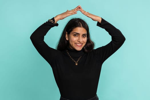 Young indian woman playing childish catching up game, feeling in safe making roof above head with hands insurance, security service. Arabian hindu girl isolated alone on blue studio background indoors