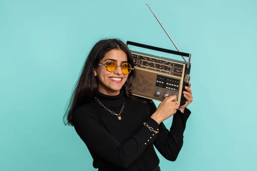 Indian woman using retro tape record player to listen music, disco dancing of favorite track, having fun, entertaining, fan of vintage technologies. Pretty hindu girl isolated on blue background
