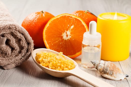 Oranges, towel, sea shell, bottle with aromatherapy oil, wooden spoon with yellow sea salt and burning candle on wooden desk. Spa products and accessories. Selective focus on salt.
