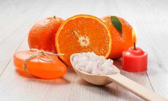 Cut orange with two whole oranges, soap, wooden spoon with white sea salt and burning red candle on wooden desk. Spa products and accessories.