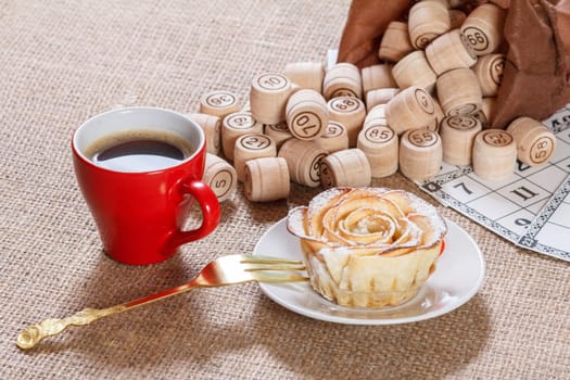 Wooden lotto barrels in brown pouch and game cards for a game in lotto with cup of coffee and homemade biscuit in the form of rose on white saucer.