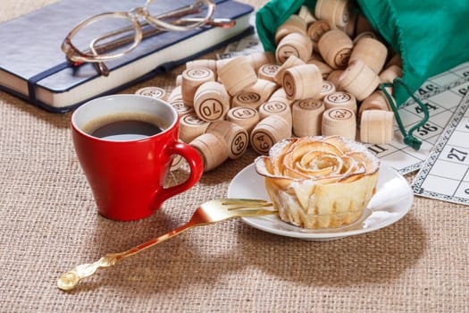 Wooden lotto barrels in green pouch and game cards for a game in lotto with notebook, glasses, red cup of coffee and homemade biscuit in the form of rose on white saucer with fork.