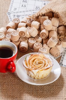 Wooden lotto barrels in pouch and game cards for a game in lotto with cup of coffee and homemade biscuit in the form of rose on white saucer. Top view.