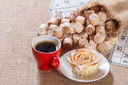 Wooden lotto barrels in pouch and game cards for a game in lotto with cup of coffee and homemade biscuit in the form of rose on white saucer. Top view.