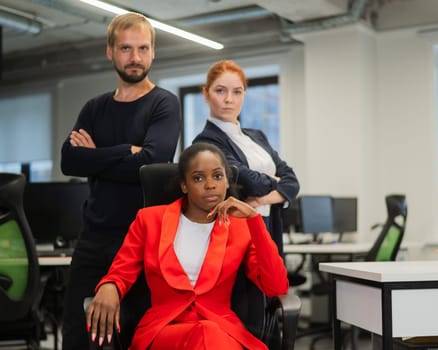 Caucasian red-haired woman, bearded Caucasian man stand behind a seated African American young woman in the office