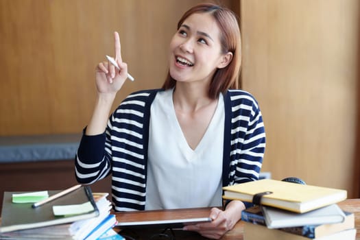 A portrait of a young Asian woman showing a smiling face and gesturing out ideas while using a tablet computer studying online at a library.