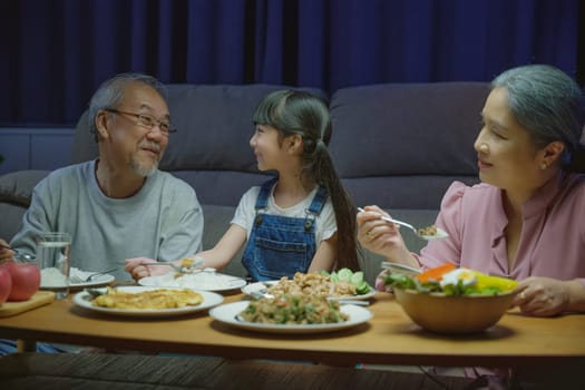 Happy Asian family grandmother grandfather and granddaughter dining on table and having fun during at home night time, senior parent and child eating food together in living room indoors together
