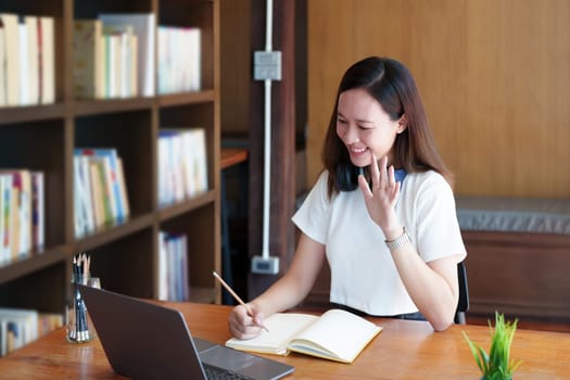 Portrait of a teenage Asian woman using a computer and notebook to study online via video conferencing on a wooden desk in library.