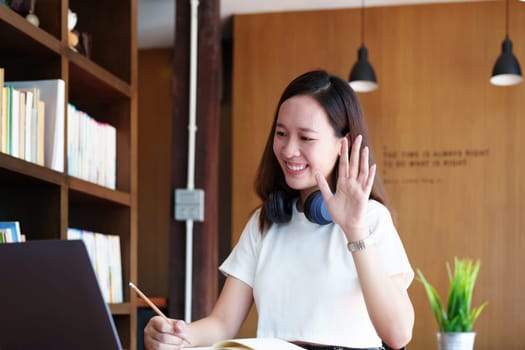 Portrait of a teenage Asian woman using a computer and notebook to study online via video conferencing on a wooden desk in library.