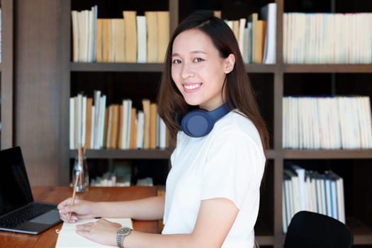 Portrait of a teenage Asian woman using a computer and notebook to study online via video conferencing on a wooden desk in library.