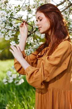 a happy, modest woman is standing in an orange dress near a flowering tree and holding her hands on the branches leans her head to them. High quality photo