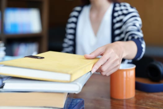 A portrait of a young Asian woman with a smiling face picking up a notebook to write down while sitting in the library reading a book.