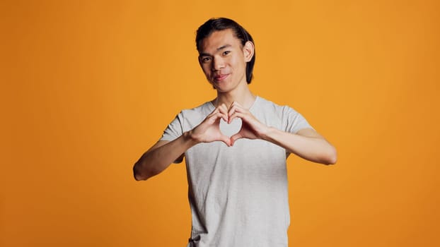 Lovely confident guy doing heart shape sign on camera, posing for vday in studio. Young adult doing romantic gesture and expressing love or beautiful feelings. Asian man showing sweet emotions.