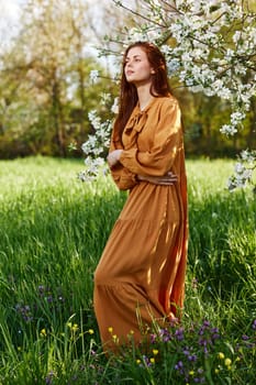 a slender, sweet woman stands in a long orange dress in the tall grass near a flowering tree and looks away with her arms crossed on her chest. High quality photo