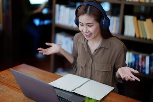 Portrait of a smiling Asian teenage girl wearing headphones and using a computer for online video conferencing in a library.