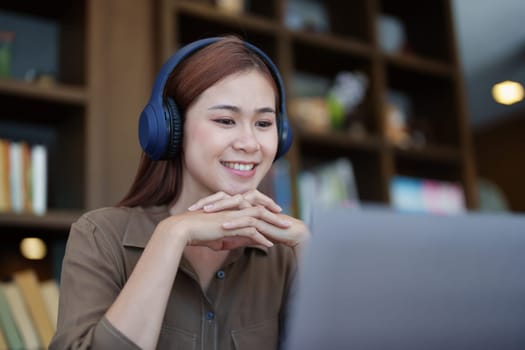 Portrait of a smiling Asian teenage girl wearing headphones and using a computer for online video conferencing in a library.