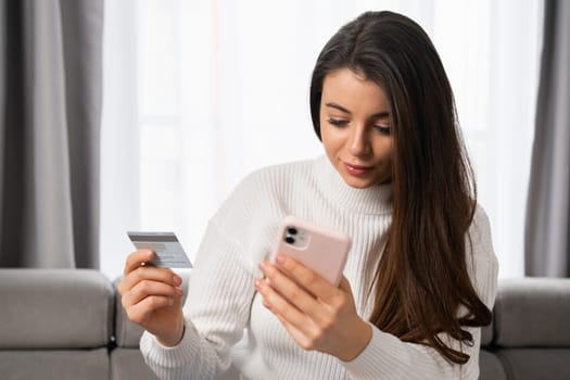 A caucasian woman with long dark hair pays by card using her mobile phone at home with a modern interior