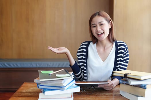 A portrait of a young Asian woman showing a smiling face and gesturing out ideas while using a tablet computer studying online at a library.