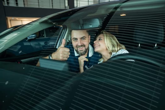 Happy caucasian couple is sitting in a new car in a car dealership. Man showing thumbs up