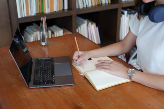 Portrait of a teenage Asian woman using a computer and notebook to study online via video conferencing on a wooden desk in library.