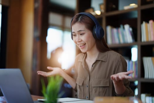 Portrait of a smiling Asian teenage girl wearing headphones and using a computer for online video conferencing in a library.