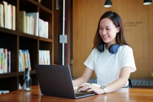 Portrait of a teenage Asian woman using a computer and notebook to study online via video conferencing on a wooden desk in library.