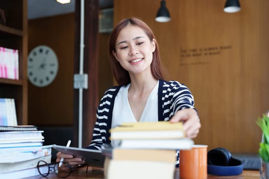 A portrait of a young Asian woman with a smiling face picking up a notebook to write down while sitting in the library reading a book.