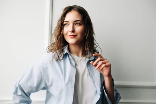 Thoughtful smiling woman with sad eyes in a blue casual shirt poses against the backdrop of a white wall at home, looks away, remembering bittersweet memories of her ex-boyfriend, playing with her hair.