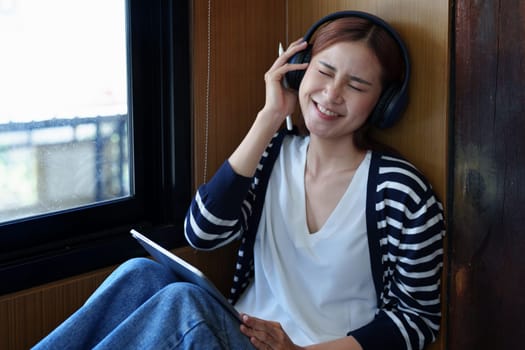 A portrait of a young Asian woman with a smiling face using a tablet computer and wearing headphones over her ears is sitting happily relaxing.