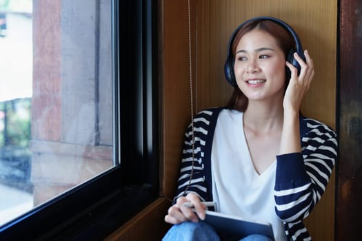 A portrait of a young Asian woman with a smiling face using a tablet computer and wearing headphones over her ears is sitting happily relaxing.