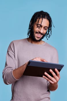 Smiling arab man typing online message on digital tablet and scrolling social media network. Happy person with cheerful face expression browsing internet and texting using gadget