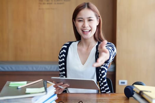 A portrait of a young Asian woman showing a smiling face and gesturing out ideas while using a tablet computer studying online at a library.