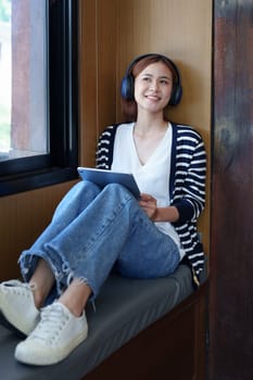 A portrait of a young Asian woman with a smiling face using a tablet computer and wearing headphones over her ears is sitting happily relaxing.