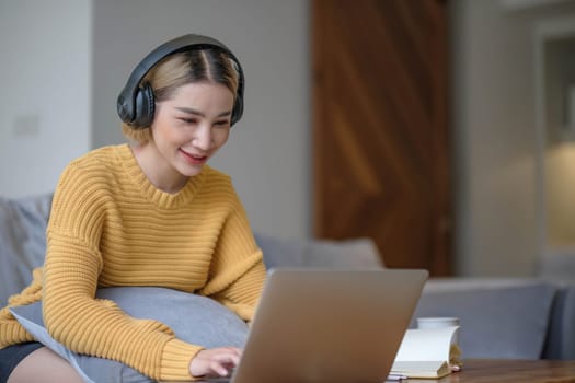 young asian woman student with headphones using laptop in a video call or online class while sitting on sofa at home.