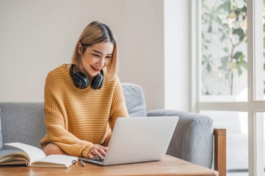 young asian woman student with headphones using laptop in a video call or online class while sitting on sofa at home.