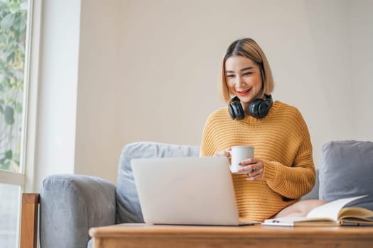 young asian woman student with headphones using laptop in a video call or online class while sitting on sofa at home.