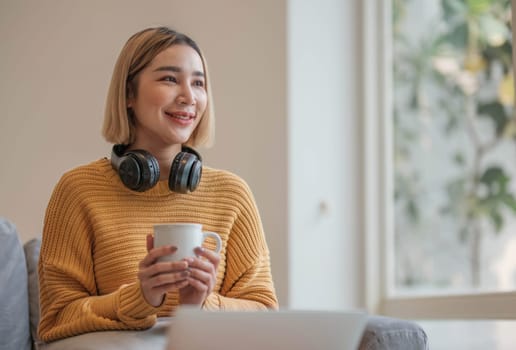 young asian woman student with headphones using laptop in a video call or online class while sitting on sofa at home.