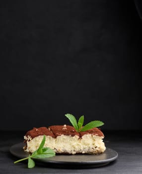 A piece of tiramisu sprinkled with cocoa, on top of a sprig of fresh mint on a black background