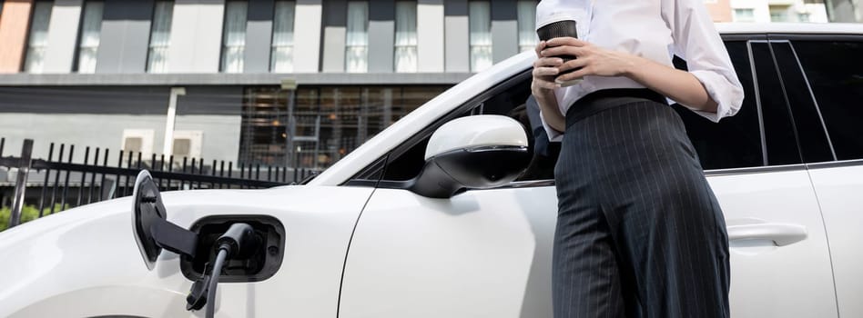 Closeup progressive suit-clad businesswoman with her electric vehicle recharge her car on public charging station in modern city with power cable plug and renewable energy-powered electric vehicle.