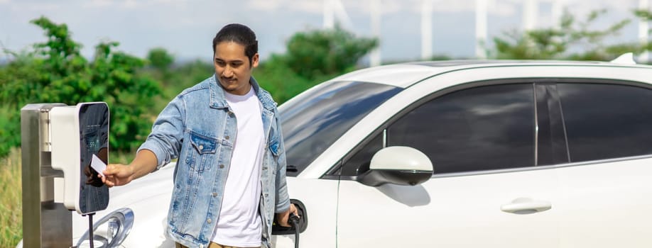 Progressive man with his electric car, EV car recharging energy from charging station on green field with wind turbine as concept of future sustainable energy. Electric vehicle with energy generator.