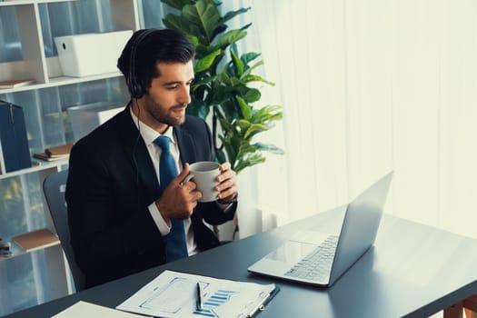 Manager of call center operator office sitting on his desk with his coffee while working on laptop. Office worker wearing headset and black suit working on customer support or telemarketing. fervent